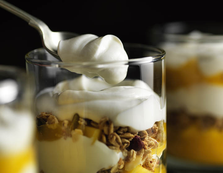 A spoon of yoghurt with a glass of yoghurt, granola and fruit