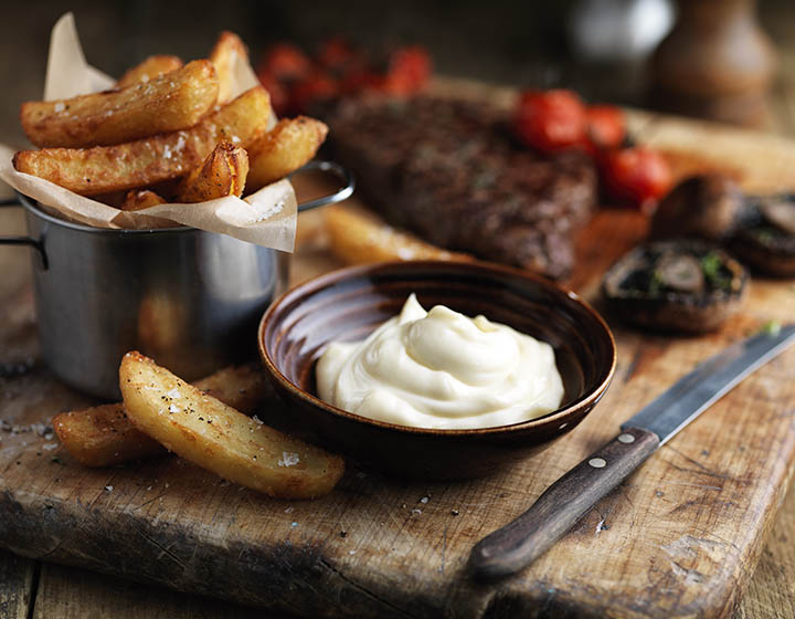A bowl of mayonnaise surrounded by steak, chips and vegetables