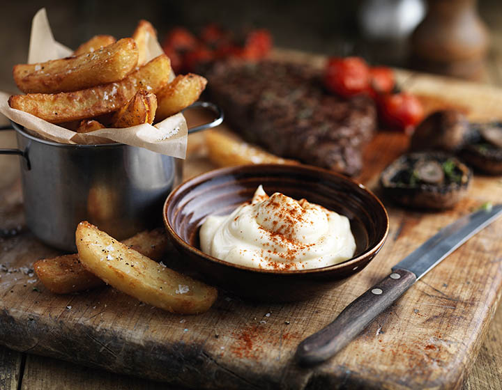 A chopping board covered in steak, tomatoes, chips and a bowl of mayonnaise