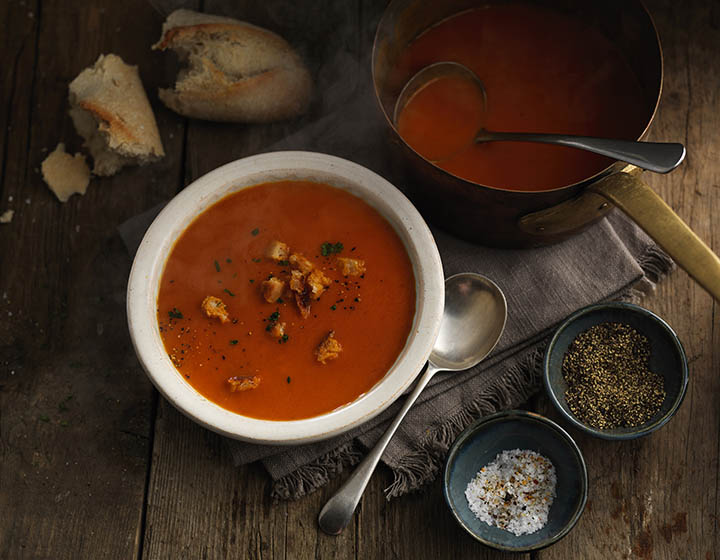 A bowl of red tomato soup with bread and smaller bowls of flavourings