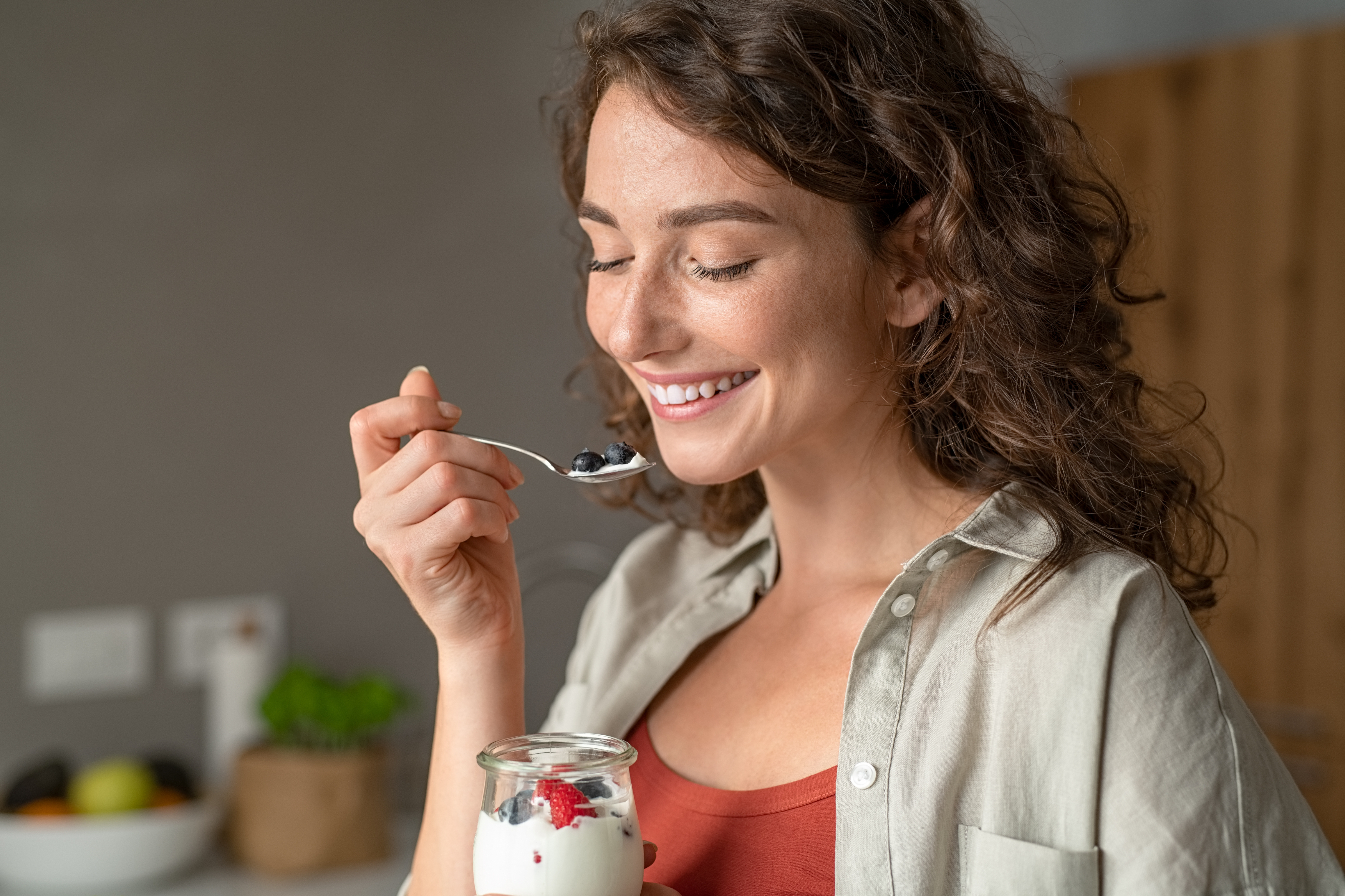 Smiling young woman having a relaxing healthy breakfast at home with fruit and yogurt. Portrait of happy natural girl holding teaspoon with yogurt and blueberries. Beautiful woman eating fresh yoghurt with berries and granola for breakfast at home.