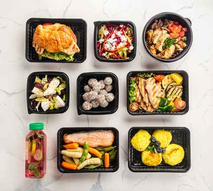 "Assorted healthy meal trays with salad, grilled chicken, salmon, vegetables, snacks, and a fruit drink on a white surface