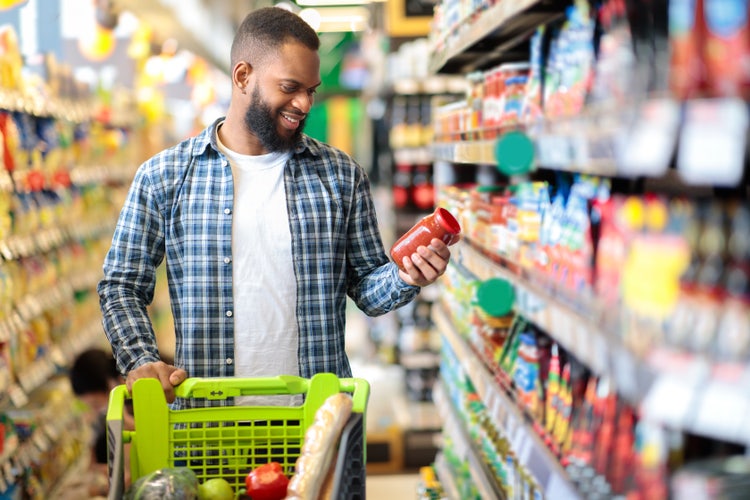 Shopper examining a jar of sauce in a grocery store aisle, with a cart full of fresh produce and bread, highlighting consumer behavior and product variety in retail environments