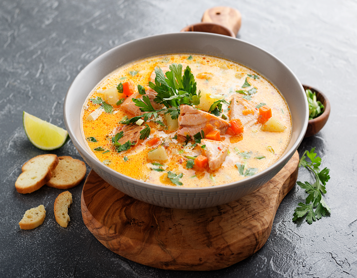 Creamy salmon and vegetable soup served in a white bowl, garnished with fresh parsley, with bread slices and lemon on a rustic table.