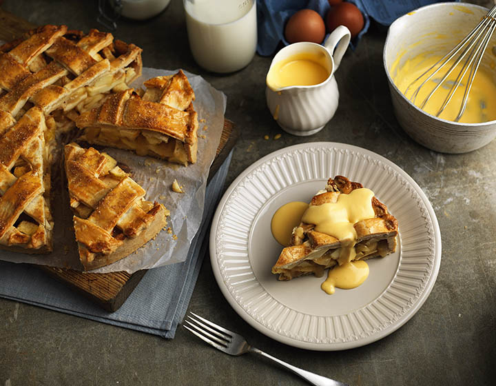 Slice of apple pie and custard next to pie, fork and jug of custard