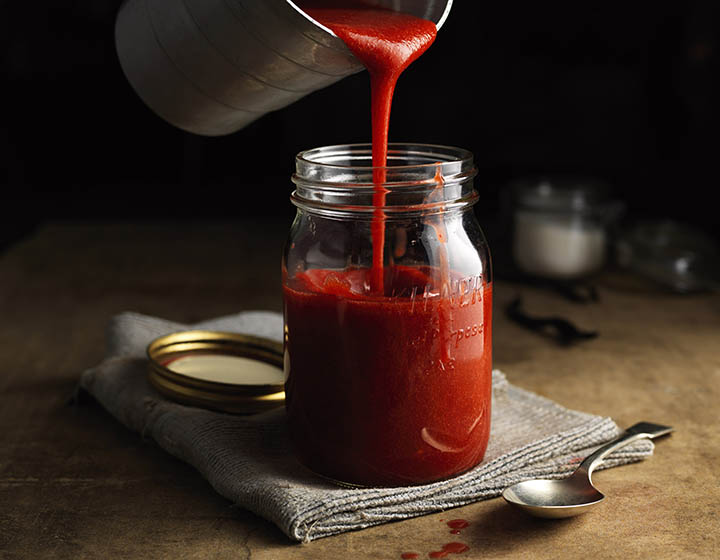 Red coulis being poured into jar, on grey tea towel, next to teaspoon