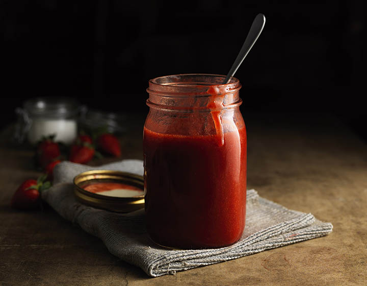 Red coulis in open jar with spoon, on grey tea towel
