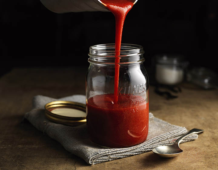 Red coulis being poured into half-filled Kilner jar, on grey tea towel, next to teaspoon
