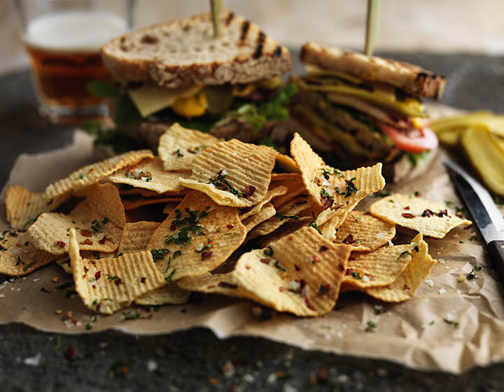 Pile of ridged crisps with sea salt and herbs on brown paper in front of sandwiches and glass of beer