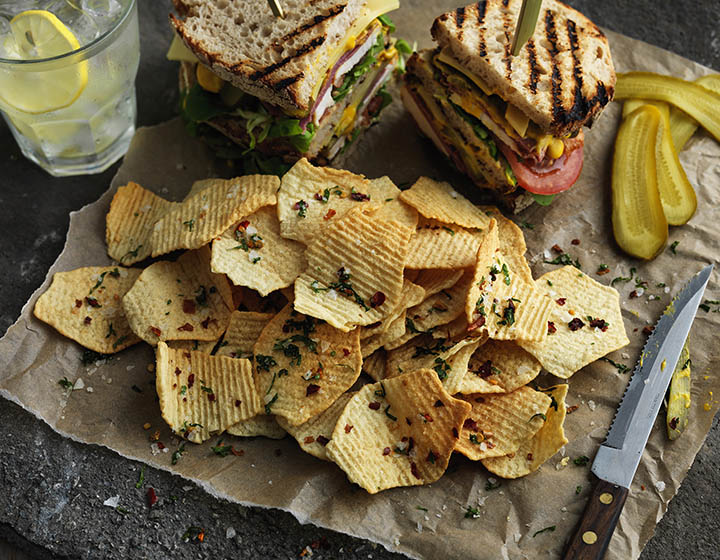 Pile of ridged crisps with sea salt and herbs on brown paper with sandwiches and knife