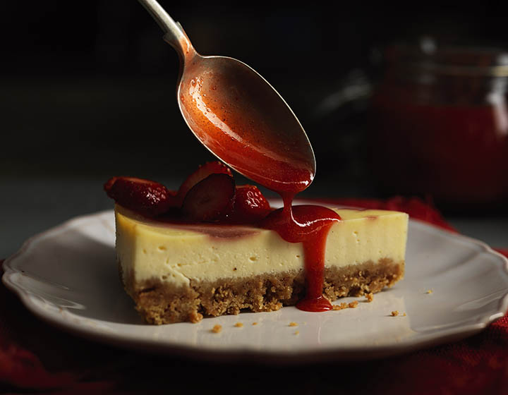 Spoon dripping red sauce onto a slice of yellow cheescake