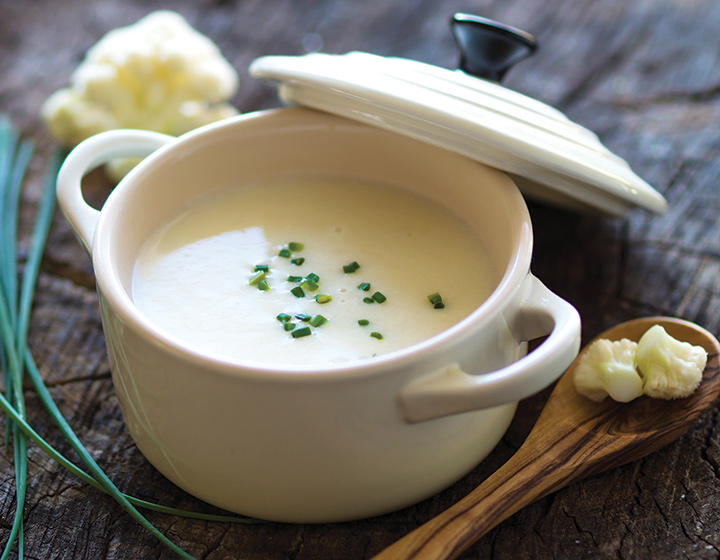 A white bowl of cauliflower soup with a white lid and a wooden spoon