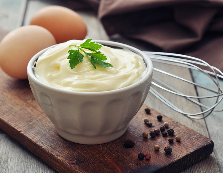 A bowl of mayonnaise with a whisk and eggs on a board