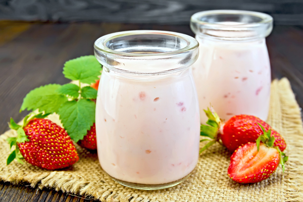 Yogurt with strawberries in two glass jar on a napkin of burlap with berries and mint on a wooden boards background