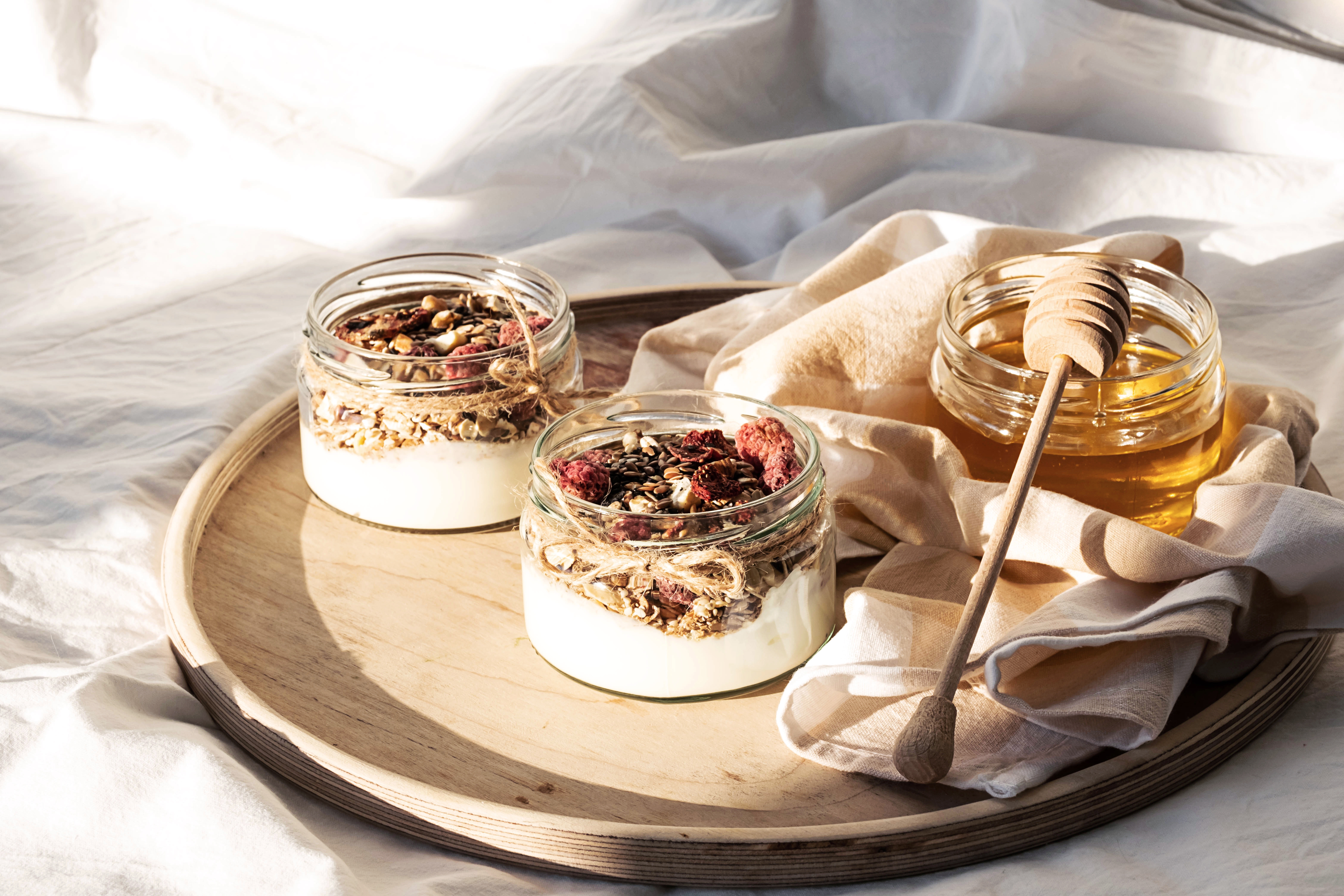 Homemade granola parfait. Quick healthy breakfast greek yogurt, honey, granola with dried berries and nuts in glass jar on wooden tray in white bedding in morning sunlight. 