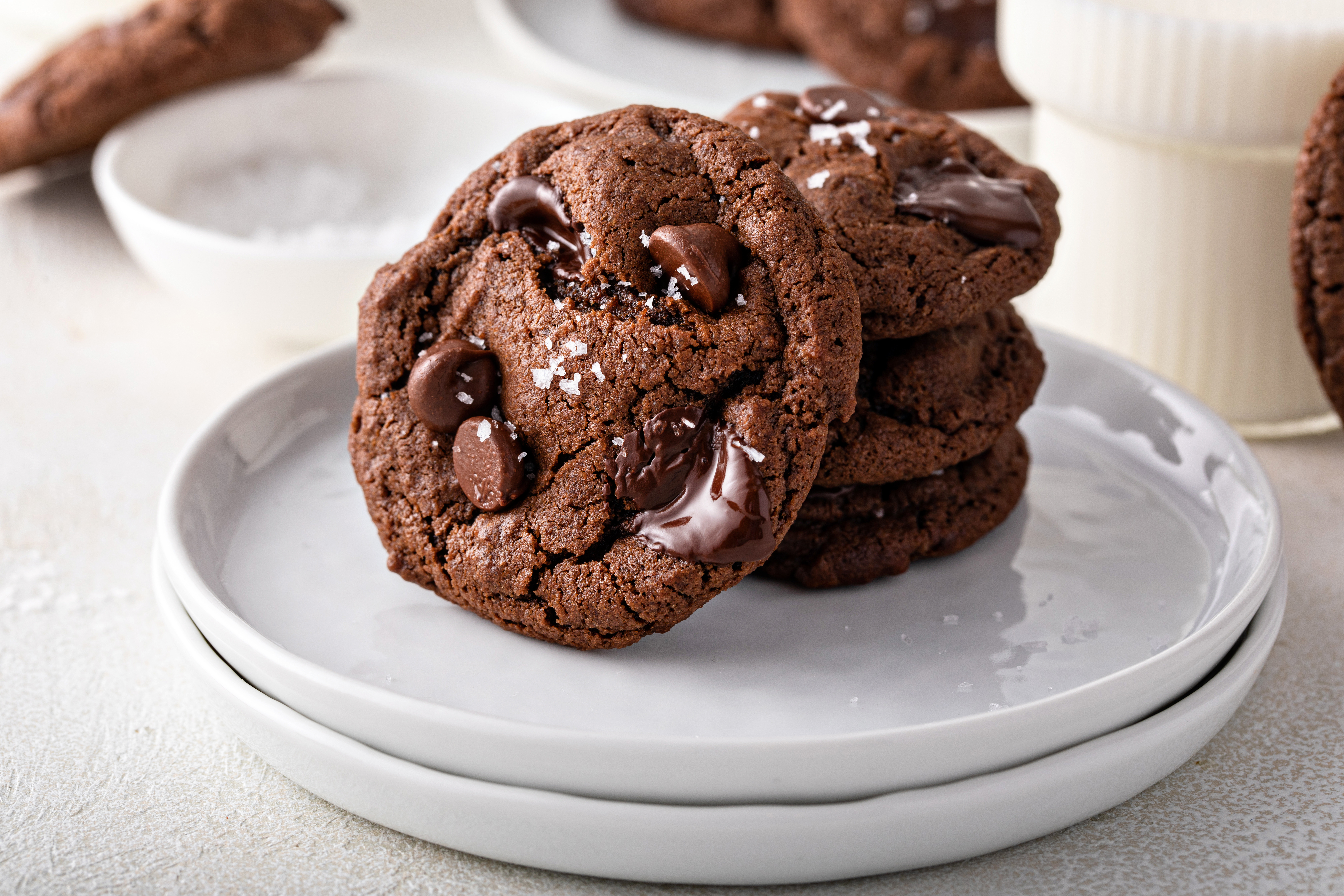Stack of three chocolate cookies with melted chocolate chips on a white plate, with a bowl and part of another cookie in the background.
