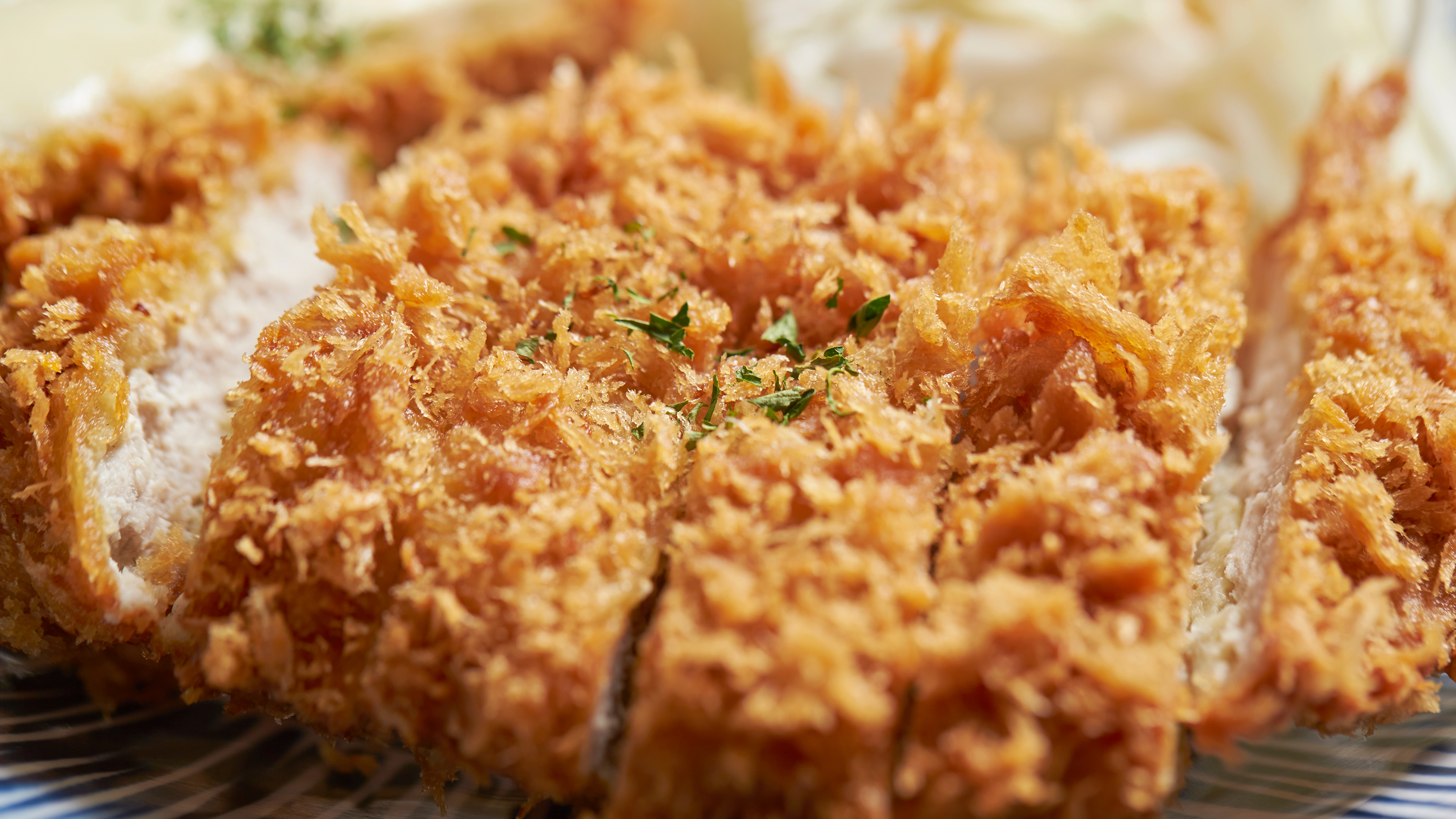 Close-up of a sliced, golden-brown breaded cutlet with crispy coating and herbs on top, with blurred food items in the background.
