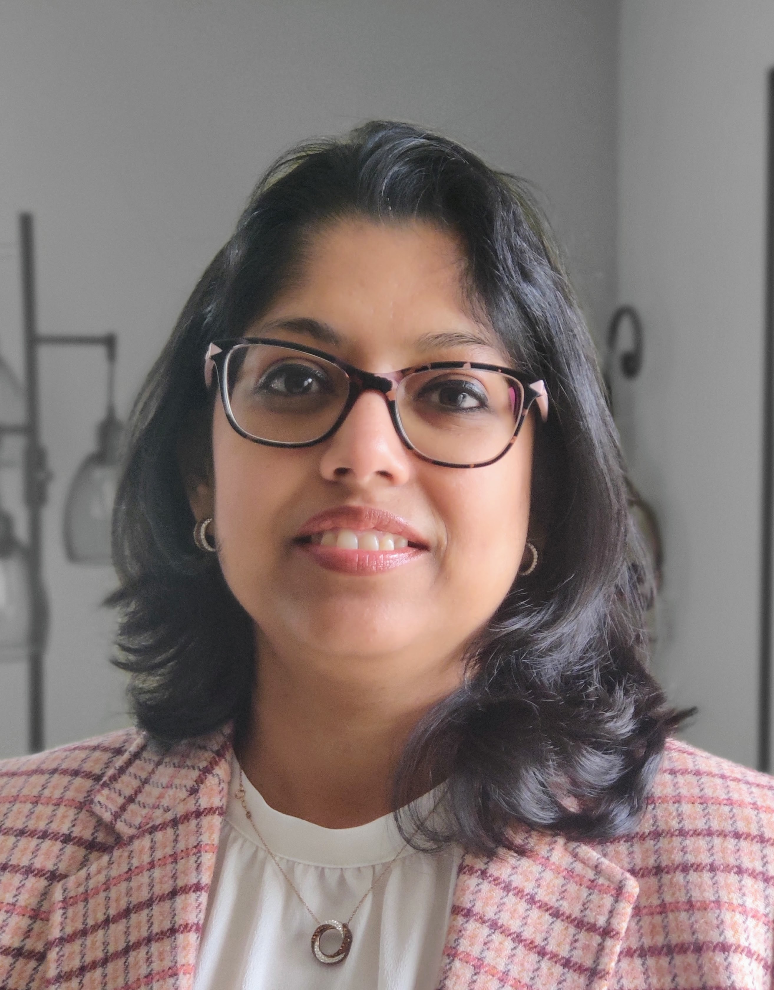 Woman with shoulder-length hair, squared glasses, a pink checkered blazer, golden earings and a white top with a necklace.