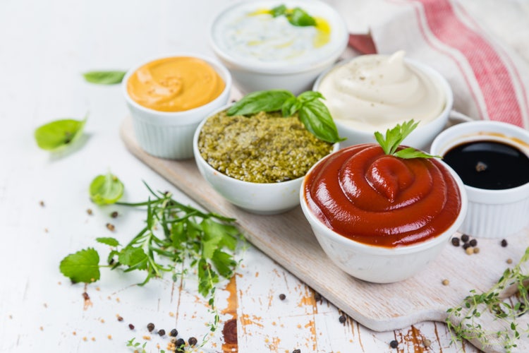 Assorted sauces and dips in white bowls on a wooden board, including ketchup, pesto, creamy and spicy condiments, garnished with herbs and peppercorns for culinary variety and flavor inspiration