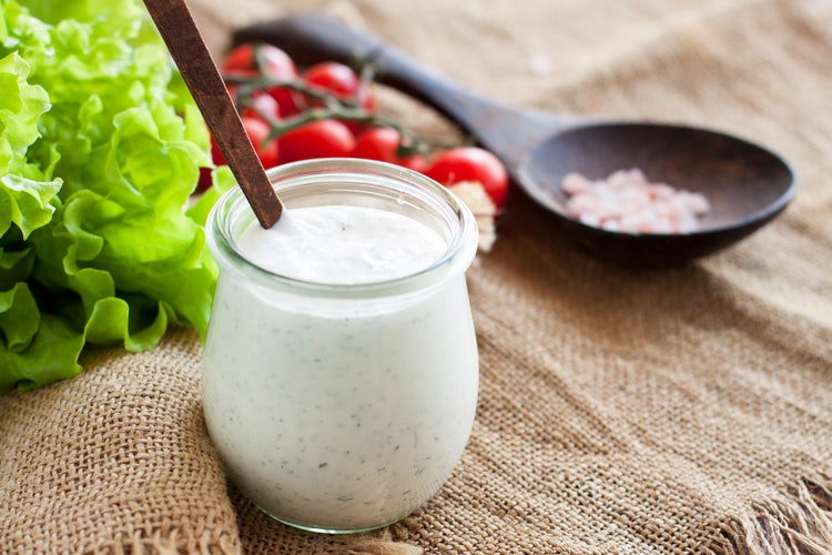 Jar of creamy ranch dressing with a wooden spoon inside, set on a burlap cloth, surrounded by fresh green lettuce, cherry tomatoes on the vine, and a wooden spoon filled with pink Himalayan salt