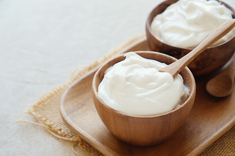 Two wooden bowls filled with creamy white yogurt, one with a wooden spoon resting inside, placed on a wooden tray over burlap fabric. The setup highlights a simple and natural presentation of a healthy dairy or plant-based food
