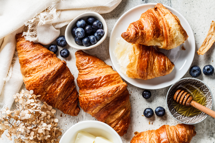 Croissants recién horneados con capas laminadas y acabado dorado, acompañados de arándanos frescos y miel