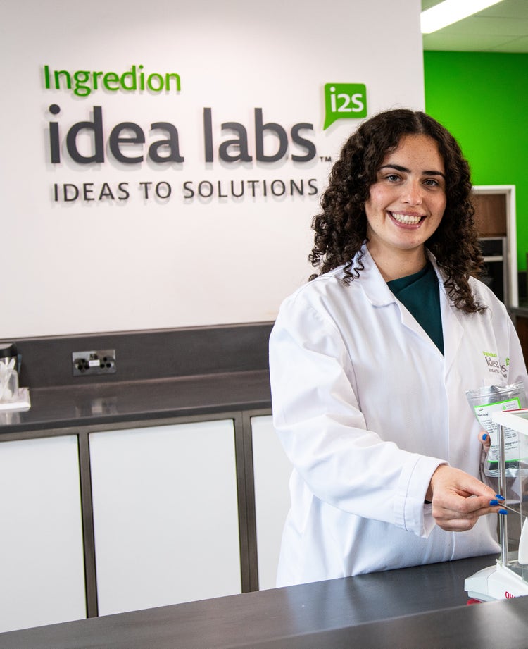 Person in a lab coat standing in front of a sign that reads 'Ingredion Idea Labs i2s IDEAS TO SOLUTIONS,' holding an object and smiling in a laboratory setting with a counter and equipment in the background