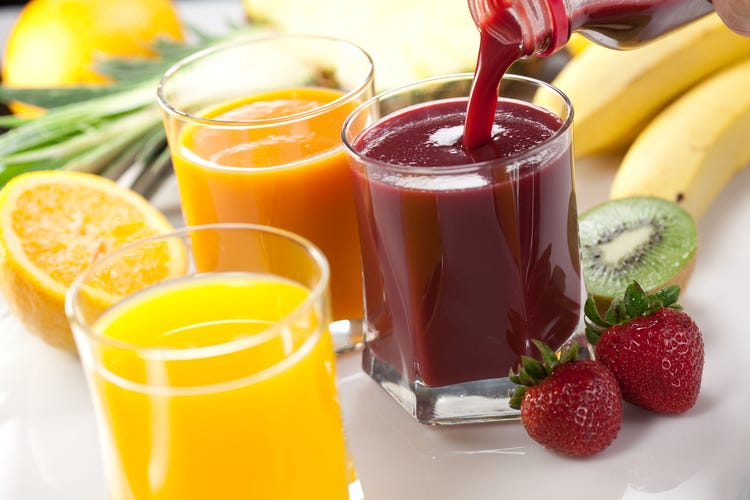 Three glasses of vibrant fruit juice—yellow, orange, and red—surrounded by fresh strawberries, kiwi, banana, and sliced orange, with red juice being poured, showcasing natural ingredients and colorful beverage variety