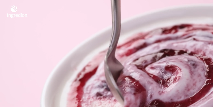 Close-up of creamy white yogurt swirled with vibrant red fruit sauce in a glass bowl, with a spoon lifting a smooth swirl against a pink background