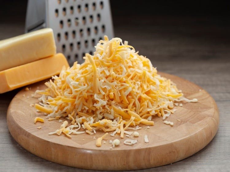 Pile of shredded cheese on a wooden cutting board with yellow and white cheese blocks behind it, and a metal box grater in the background. The image highlights the cheese grating process and variety of cheese types.