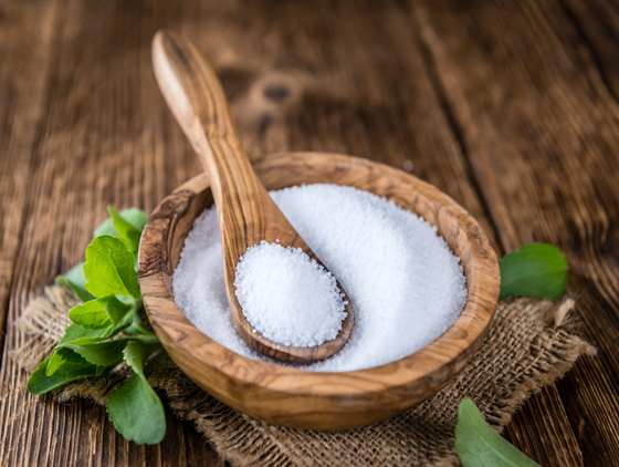 Wooden bowl filled with white granulated sweetener and a wooden spoon resting on top, placed on a rustic burlap cloth with fresh green leaves on a wooden table
