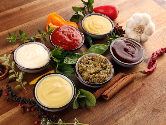 Assorted sauces in small bowls on a wooden surface, including creamy white, yellow, red, brown, and green varieties, surrounded by fresh herbs, garlic, peppers, cinnamon sticks, and dried chili