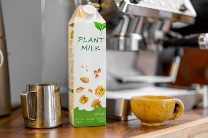 Carton of plant milk labeled 'SUGAR FREE,' 'LACTOSE FREE,' and 'GLUTEN FREE,' placed on a wooden surface next to a metal milk frothing pitcher and a yellow ceramic cup, with an espresso machine in the background. The carton features images of almonds and walnuts and has a 900 ml capacity