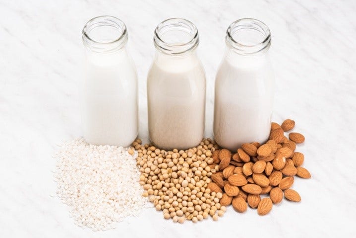 Three glass bottles of plant-based milk placed in front of their ingredients: rice, soybeans, and almonds. The image showcases non-dairy milk alternatives and their natural sources
