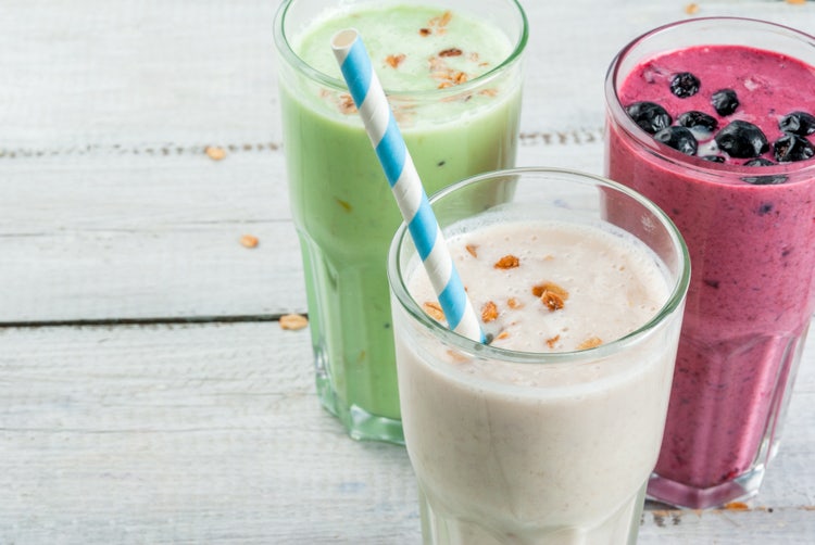 Three tall glasses of colorful smoothies on a light wooden surface: a creamy white smoothie with granola and striped straw in the foreground, a green smoothie with granola to the left, and a vibrant purple smoothie garnished with blueberries to the right, showcasing fresh ingredients and healthy drink options
