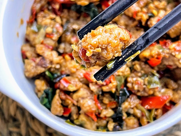 Close-up of stir-fried chicken and vegetables, including red bell peppers and greens, held by chopsticks above a white bowl.