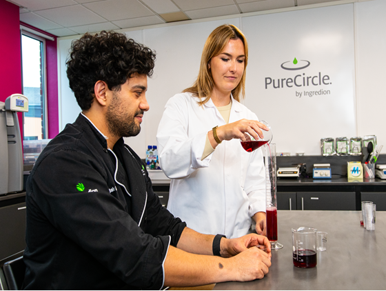 Two people in a laboratory setting working with a tall graduated cylinder filled with a red liquid, with additional measuring cups on the counter and the PureCircle by Ingredion logo visible on the wall