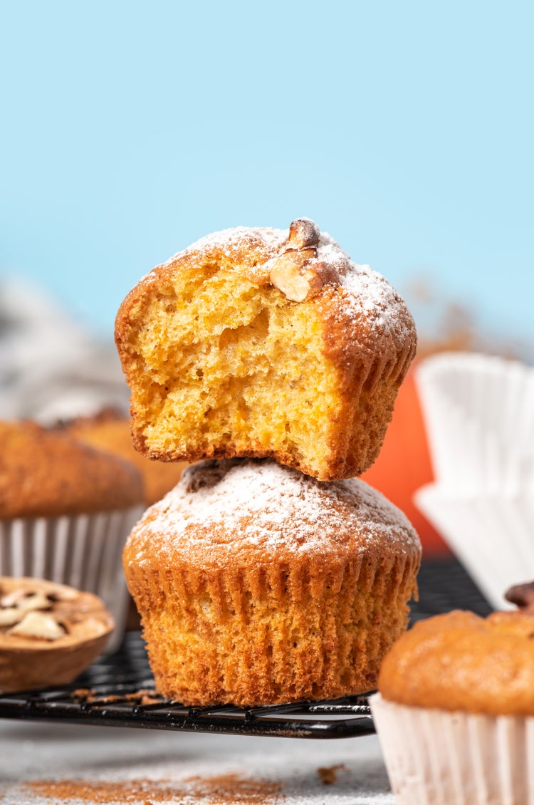 Two golden-brown muffins stacked on a cooling rack, with the top muffin partially bitten and dusted with powdered sugar, surrounded by other muffins and baking cups against a light blue background