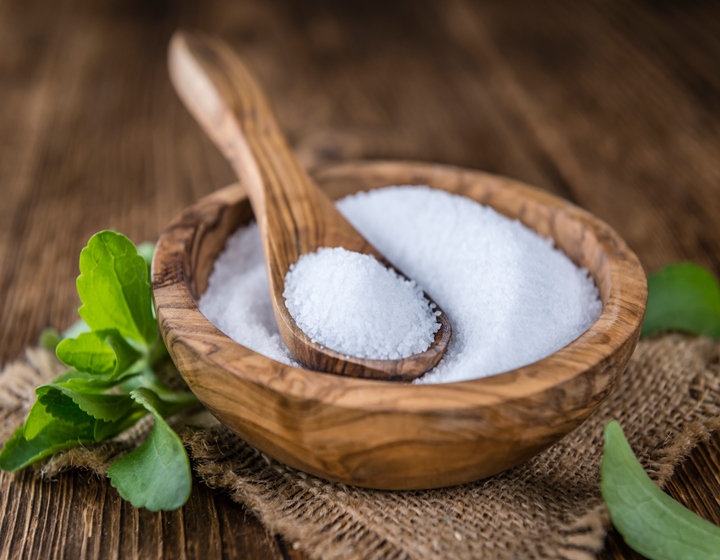 Wooden bowl filled with granulated white sugar and a wooden spoon on a rustic table, with fresh green leaves for decoration