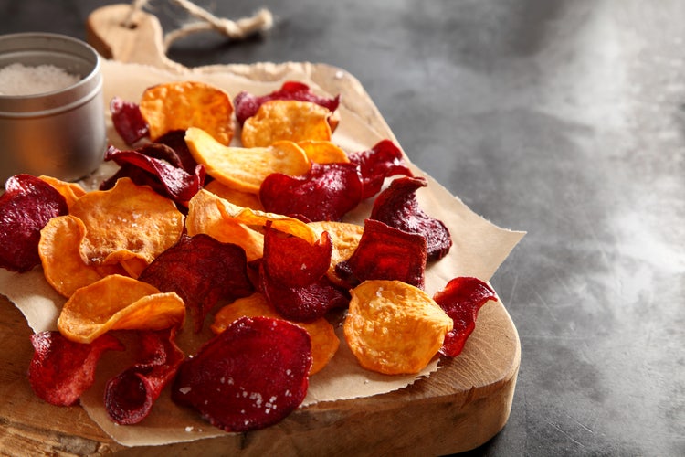 Colorful red and orange salty chips on parchment paper over a wooden cutting board, with a small metal container of coarse salt nearby.