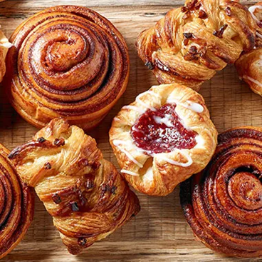 Assorted flaky pastries on a wooden surface, including cinnamon rolls and a pastry topped with fruit jam and icing