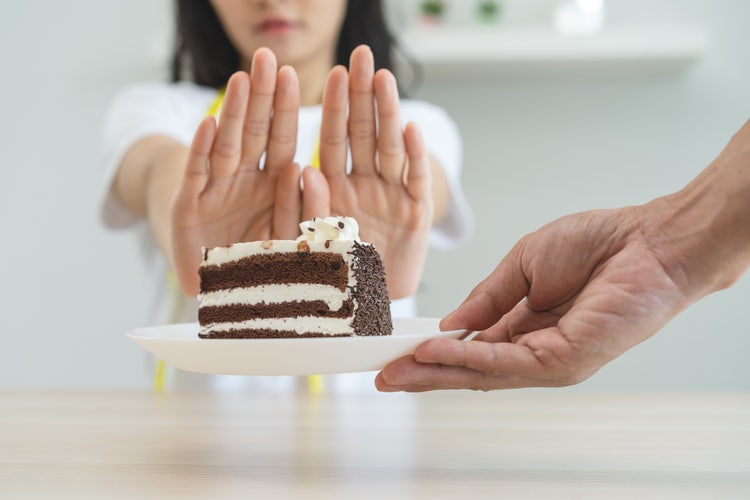 Person holding up hands to refuse a plate with a slice of chocolate layer cake being offered