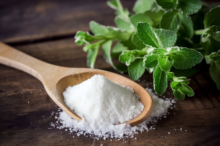 Wooden spoon filled with white granulated stevia powder on a rustic wooden surface, with fresh green stevia leaves in the background