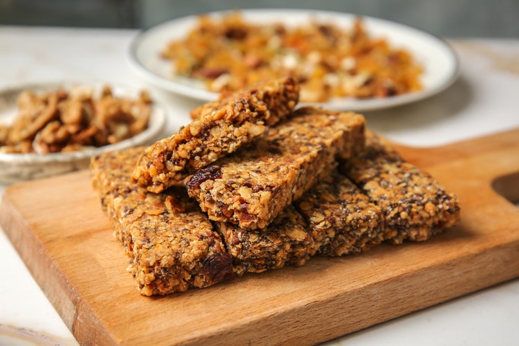 Stack of granola bars with oats, nuts, and dried fruits on a wooden board, with bowls of trail mix and nuts in the background.
