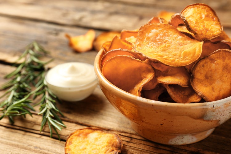 A rustic bowl of golden-brown sweet potato chips beside a small dish of white dip and a sprig of rosemary on a wooden surface.