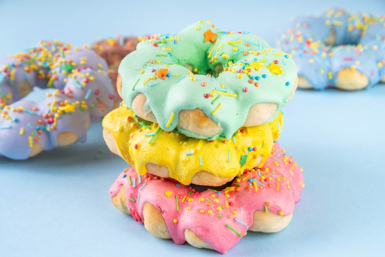 Stack of three colorful mochi donuts with pastel icing in green, yellow, and pink, topped with rainbow sprinkles, with more donuts in the background on a light blue surface