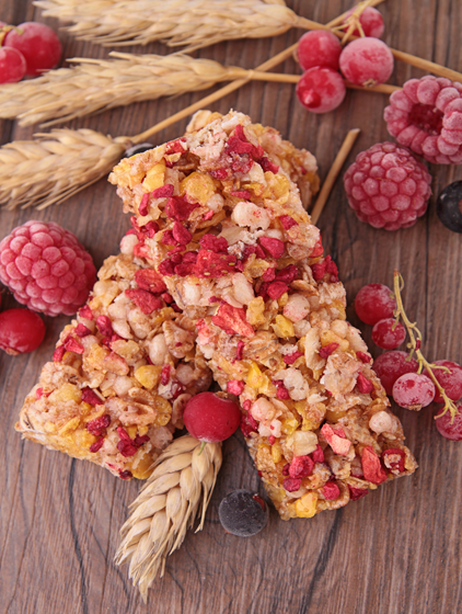 Two granola bars topped with dried fruits, mainly red berries and yellow pieces, placed on a wooden surface. Surrounding them are wheat stalks and frozen fruits such as raspberries, currants, and blueberries
