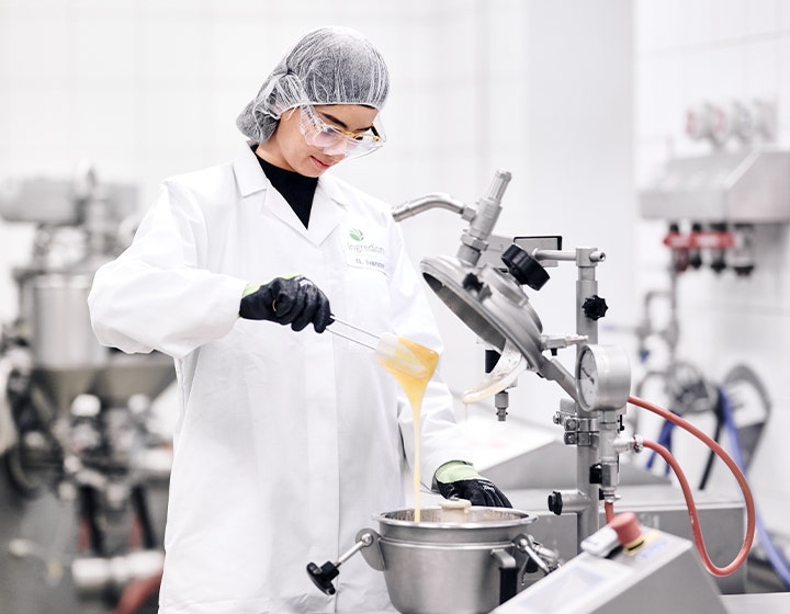 A scientist in a lab coat and hairnet pouring a viscous ingredient into machinery