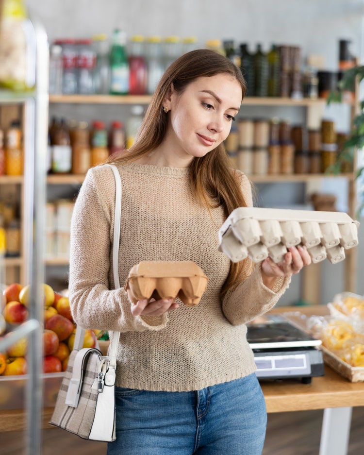 Persona en tienda de abarrotes sosteniendo dos cartones de huevos, comparando productos frescos en sección de alimentos, concepto de compra responsable y selección de ingredientes.
