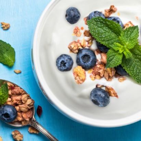 Bowl of creamy yogurt topped with fresh blueberries, crunchy granola, and mint leaves on a bright blue background